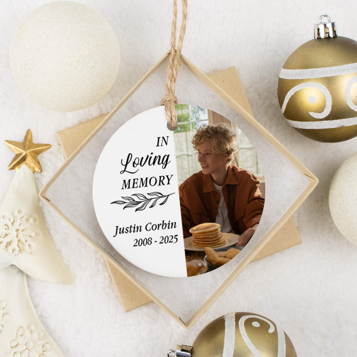 A personalized round hanging memorial ornament displayed inside a square-shaped gift box. The ornament has a white background with black text reading 'In Loving Memory' and the name 'Justin Corbin 2008-2025,' plus a printed photo of a man in a brown jacket beside a stack of pancakes.
