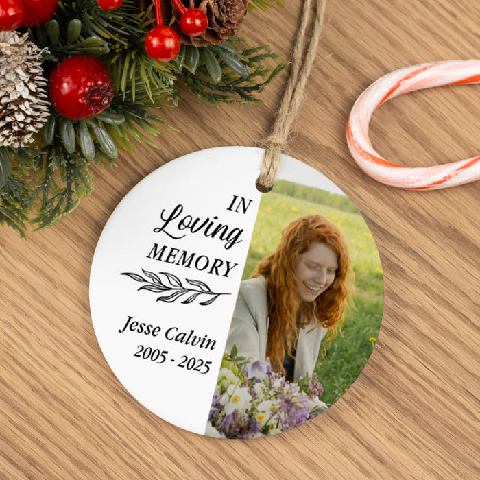 A round hanging memorial ornament on a wooden surface, decorated with holiday greenery, red berries, and a candy cane. The ornament is split into two halves: one side is white with the text 'IN Loving MEMORY Jesse Calvin 2005 - 2025' and the other side shows a photo of a smiling red-haired woman holding flowers.