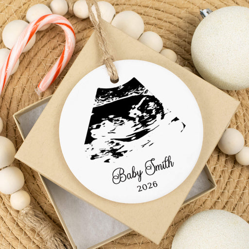A round white personalized baby keepsake ornament with a black ultrasound print and the text 'Baby Smith 2026,' hanging from a kraft paper tag on a gift box. The ornament is styled on a woven surface with candy canes, wooden beads, and white ornaments.