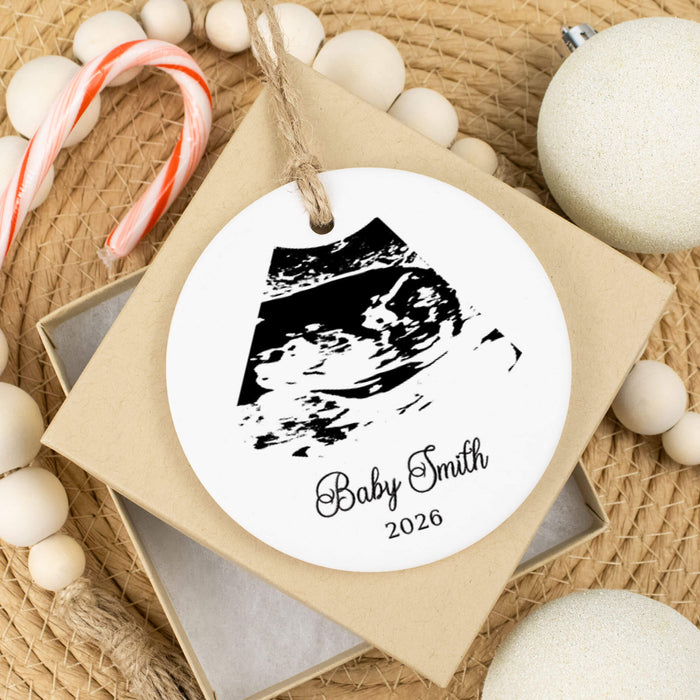 A round white personalized baby keepsake ornament with a black ultrasound print and the text 'Baby Smith 2026,' hanging from a kraft paper tag on a gift box. The ornament is styled on a woven surface with candy canes, wooden beads, and white ornaments.