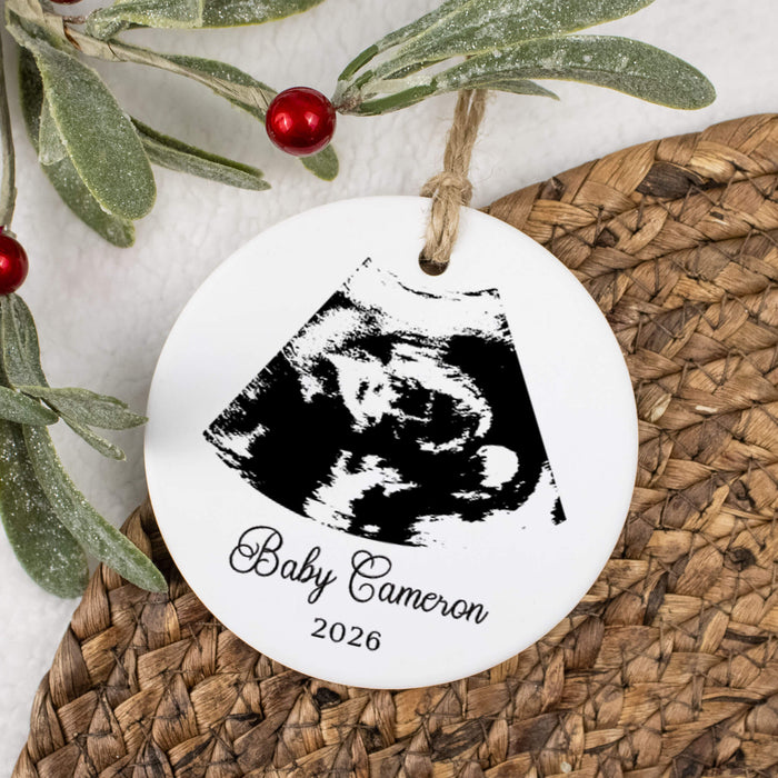 A round white personalized Christmas ornament hanging from a twine string, printed with a black ultrasound image and the text 'Baby Cameron 2026'. The ornament is styled on a woven wicker mat with green leaves and red berries in the background.