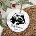 A round white personalized Christmas ornament hanging from a twine string, printed with a black ultrasound image and the text 'Baby Cameron 2026'. The ornament is styled on a woven wicker mat with green leaves and red berries in the background.