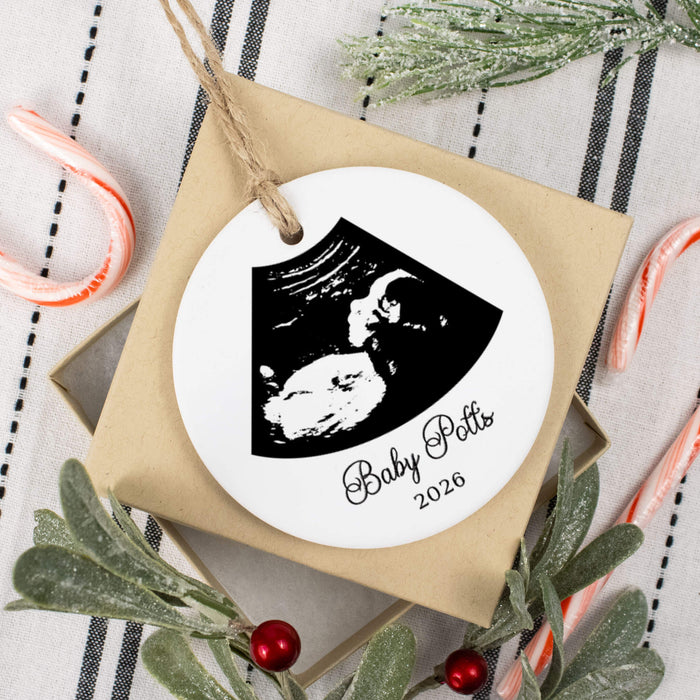 A personalized round white Christmas ornament with a black ultrasound photo and the text 'Baby Potts 2026', hanging from a kraft paper gift bag with a jute loop. The bag sits on a striped fabric surface with holiday greenery, candy canes, and red berries around it.