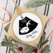 A personalized round white Christmas ornament with a black ultrasound photo and the text 'Baby Potts 2026', hanging from a kraft paper gift bag with a jute loop. The bag sits on a striped fabric surface with holiday greenery, candy canes, and red berries around it.