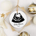A personalized round white Christmas ornament with a black ultrasound graphic and the text 'Baby Michaels 2027', hanging in a kraft paper square box. The ornament is styled on a white textured surface with gold and white holiday ornaments and decorative props.