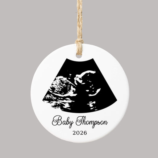 A round white ceramic ornament with a hanging loop, featuring a black ultrasound scan graphic and the personalized text 'Baby Thompson 2026' on a plain gray background.