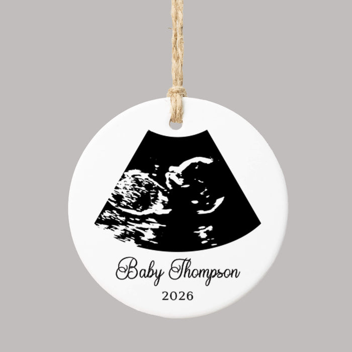 A round white ceramic ornament with a hanging loop, featuring a black ultrasound scan graphic and the personalized text 'Baby Thompson 2026' on a plain gray background.