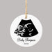 A round white ceramic ornament with a hanging loop, featuring a black ultrasound scan graphic and the personalized text 'Baby Thompson 2026' on a plain gray background.