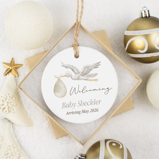 Decorative ornament with baby announcement text, surrounded by Christmas ornaments on a light background