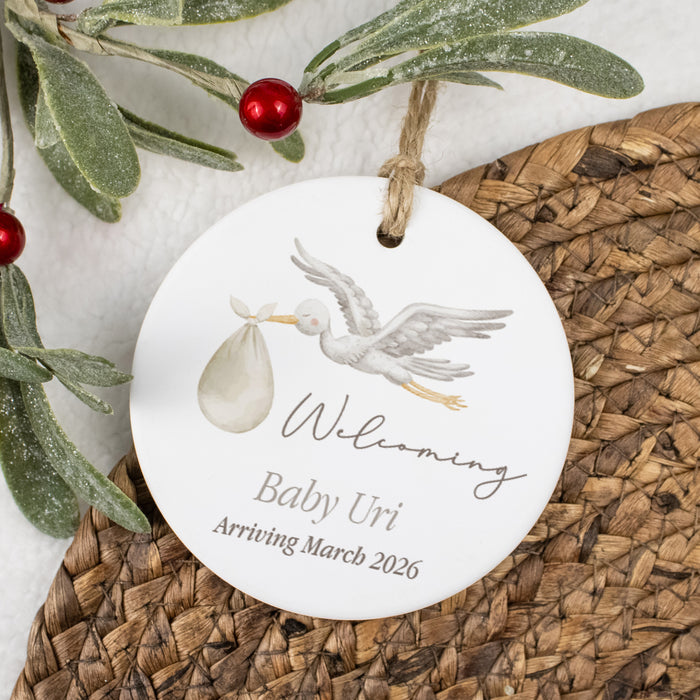 Round ceramic ornament with stork and baby design, text 'Welcoming Baby Uri Arriving March 2026', on a woven surface with mistletoe.