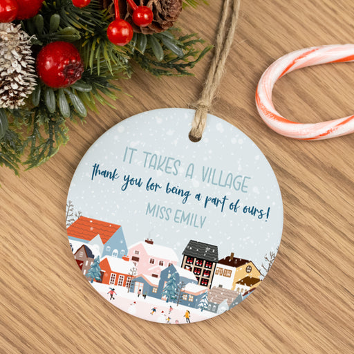 Round Christmas ornament with village scene and personalized text on a wooden surface with festive decorations for a nanny, teacher or babysitter