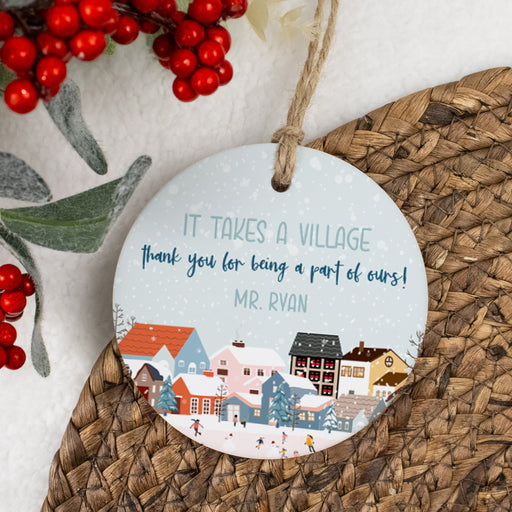it takes a village thank you for being a part of ours round ceramic ornament with personalized name laying on a woven placemat with holly berries