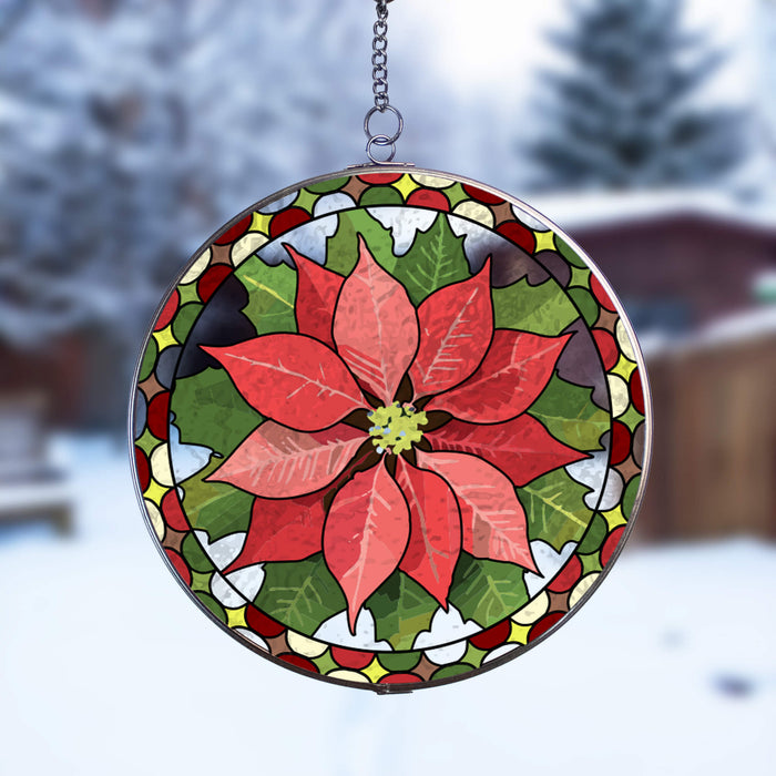 A circular stained-glass holiday suncatcher featuring a red poinsettia flower with green leaves and a decorative border, suspended by a metal chain against a snowy outdoor background.