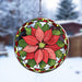 A circular stained-glass holiday suncatcher featuring a red poinsettia flower with green leaves and a decorative border, suspended by a metal chain against a snowy outdoor background.