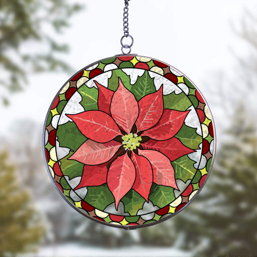 A circular stained-glass suncatcher featuring a red poinsettia flower with green leaves, hanging by a metal chain against a blurred outdoor winter background.