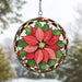 A circular stained-glass suncatcher featuring a red poinsettia flower with green leaves, hanging by a metal chain against a blurred outdoor winter background.