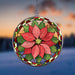 A circular stained-glass-style hanging suncatcher featuring a large red poinsettia flower over green leaves, suspended by a metal chain against a blurred outdoor sunset background.