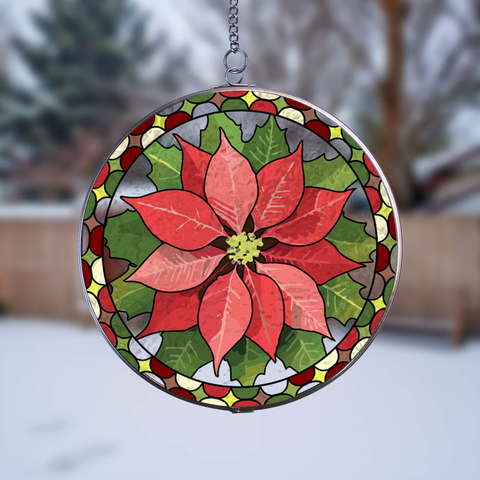 A circular stained-glass-style suncatcher featuring a large red poinsettia flower with green leaves and a decorative border, suspended outdoors against a blurred snowy background.