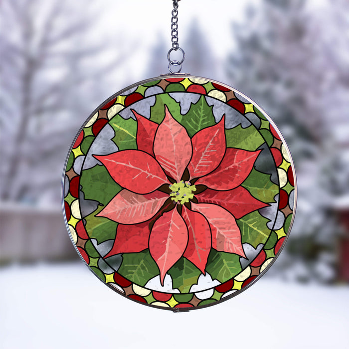 A circular stained-glass style hanging suncatcher featuring a large red poinsettia flower with green leaves and a decorative border, suspended by a chain against a blurred snowy outdoor background.