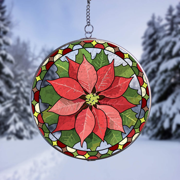 A circular stained-glass suncatcher featuring a poinsettia flower design, suspended by a metal chain against a snowy outdoor background with blurred evergreen trees.