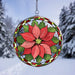 A circular stained-glass suncatcher featuring a poinsettia flower design, suspended by a metal chain against a snowy outdoor background with blurred evergreen trees.
