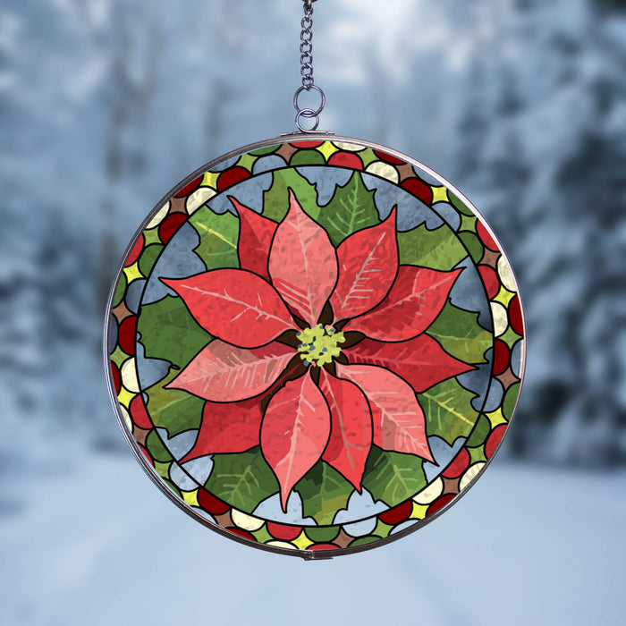 A circular stained-glass suncatcher featuring a red poinsettia flower and green leaves, suspended by a short chain against a blurred snowy outdoor background.