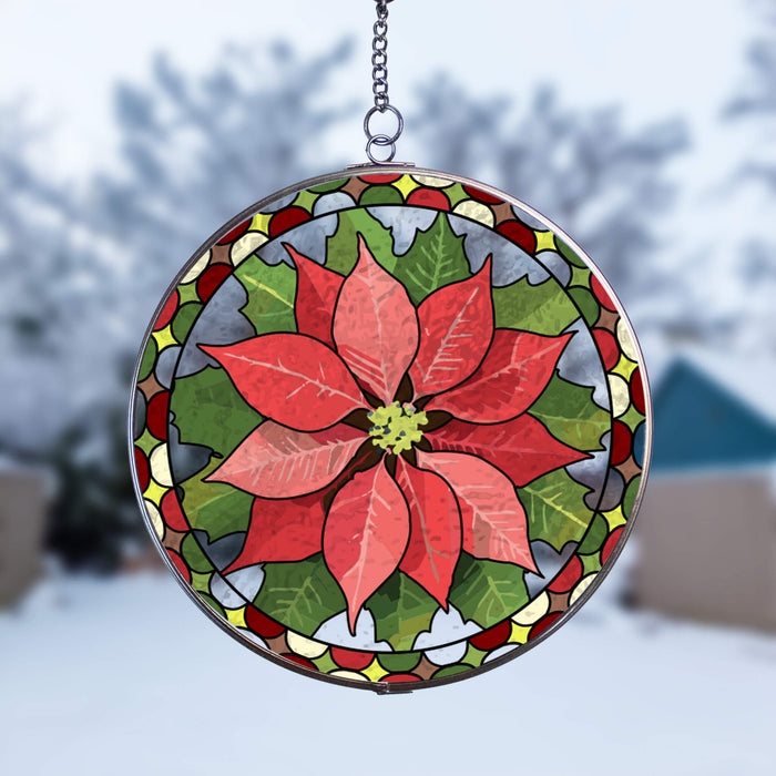 A circular stained-glass suncatcher featuring a large red poinsettia flower with green leaves and a decorative border, suspended by a metal chain against a blurred outdoor winter background.