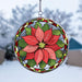 A circular stained-glass suncatcher featuring a large red poinsettia flower with green leaves and a decorative border, suspended by a metal chain against a blurred outdoor winter background.