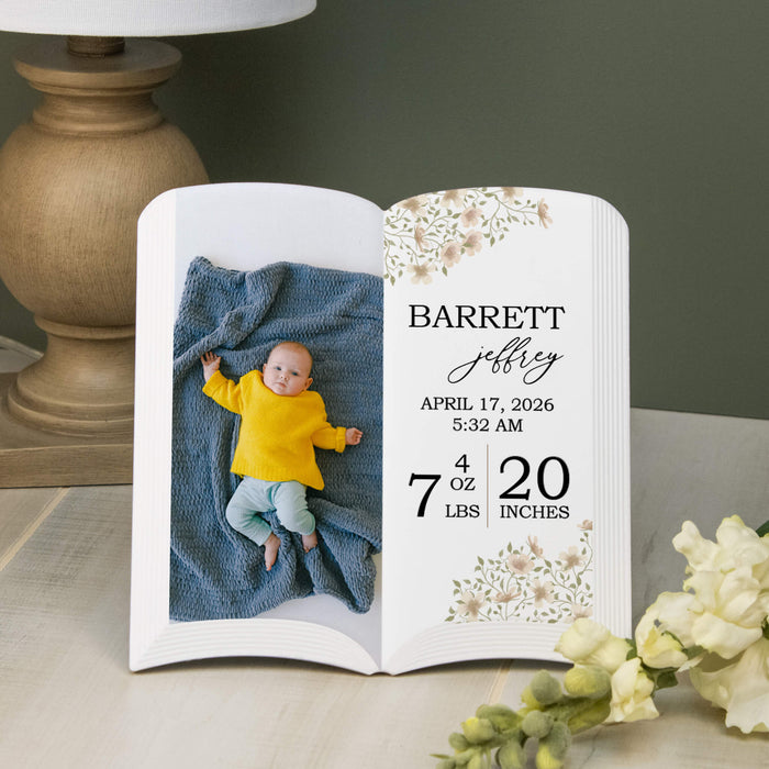 Personalized baby book with photo of a baby in a yellow shirt and blue blanket, on a table with a lamp and flowers.
