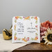 Personalized ceramic book pages with floral design and religious text on a wooden surface with flowers and a gold bird ornament.