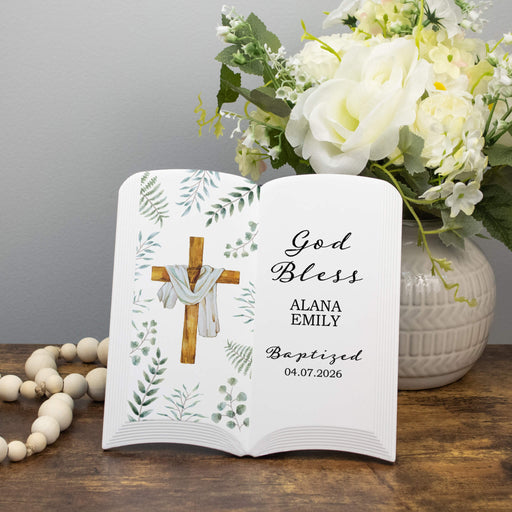 Personalized book with cross design and floral elements, placed on a wooden surface with flowers and beads.