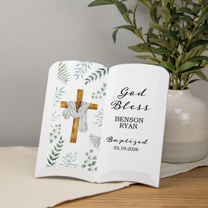 custom religious keepsake ceramic book with name, event and date beside a cross with greenery