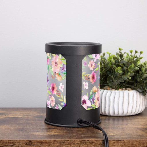 back view of a black wax warmer lamp with pink floral design on a wooden surface next to a plant.