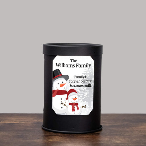 A black cylindrical wax melt lamp with a custom printed label featuring two snowmen and the text 'The Williams Family' and 'Family is forever because love never melts', set on a wooden surface against a plain gray background.