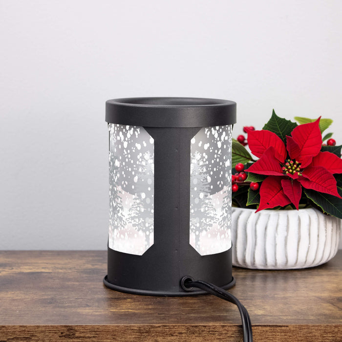 A black cylindrical electric wax warmer with frosted snowflake-patterned panels, shown on a wooden surface next to a potted red poinsettia.