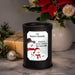 A black cylindrical wax warmer with a custom printed label reading 'The Barret Family' and a winter-themed snowman illustration, styled on a marble surface with holiday decor including poinsettias, a wrapped gift, pine branches, and a vase in the background.