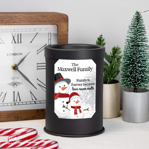A black cylindrical wax melt lamp with a custom printed snowman illustration and the text 'The Maxwell Family, Family is forever because love never melts' sits on a tabletop. The scene includes a wooden clock, candy canes, small potted plants, and a miniature Christmas tree in the background.