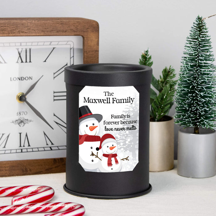 A black cylindrical wax melt lamp with a custom printed snowman illustration and the text 'The Maxwell Family, Family is forever because love never melts' sits on a tabletop. The scene includes a wooden clock, candy canes, small potted plants, and a miniature Christmas tree in the background.