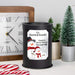 A black cylindrical wax melt lamp with a custom printed snowman illustration and the text 'The Maxwell Family, Family is forever because love never melts' sits on a tabletop. The scene includes a wooden clock, candy canes, small potted plants, and a miniature Christmas tree in the background.