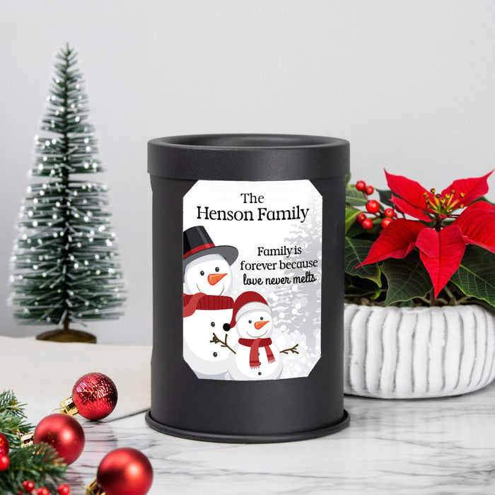 A black cylindrical wax warmer lamp with a custom printed label featuring snowman illustrations and the text 'The Henson Family, Family is forever because love never melts.' The lamp is styled on a marble surface with a small Christmas tree, red ornaments, and a potted poinsettia in the background.