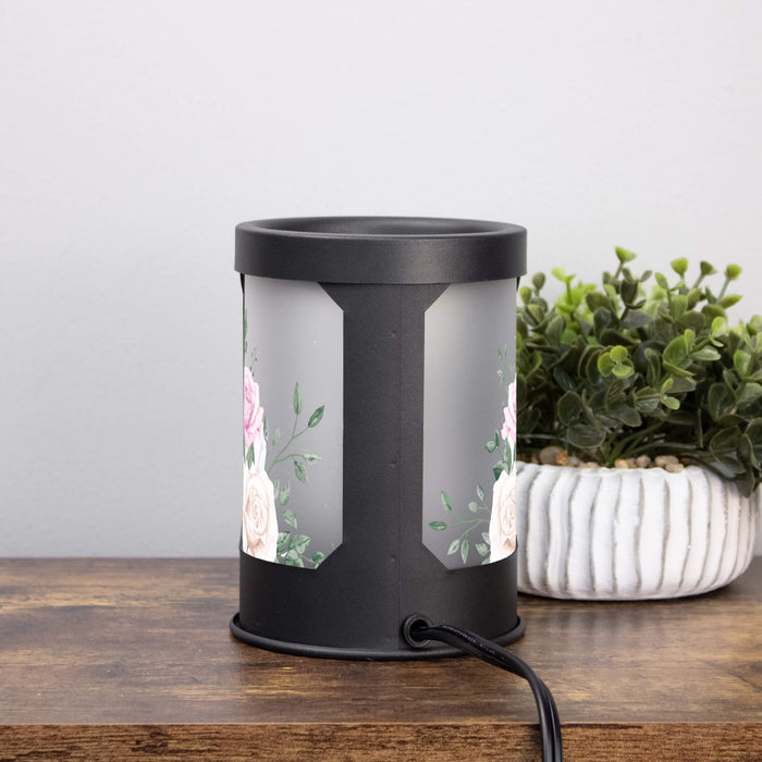Black cylindrical wax melt lamp with floral design on a wooden surface next to a plant.