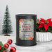 A black cylindrical wax warmer lamp with a decorative holiday label reading 'Wishing You Peace & Joy This Holiday Season' and 'The Queens,' styled on a marble surface with a small Christmas tree, red ornaments, and a poinsettia in the background.