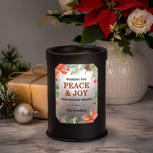 A black cylindrical wax warmer lamp with a festive holiday label reading 'Wishing You Peace & Joy This Holiday Season' and 'The Gordons,' styled on a marble surface with poinsettias, a wrapped gift, pine branches, and a white vase in the background.