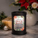 A black cylindrical wax warmer lamp with a festive holiday label reading 'Wishing You Peace & Joy This Holiday Season' and 'The Gordons,' styled on a marble surface with poinsettias, a wrapped gift, pine branches, and a white vase in the background.