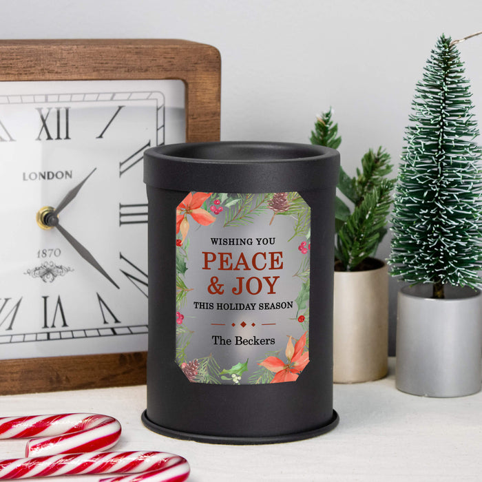 A black cylindrical wax melt lamp with a personalized holiday label reading 'Wishing You PEACE & JOY This Holiday Season The Beckers,' styled on a tabletop with a clock, small potted trees, and candy canes.