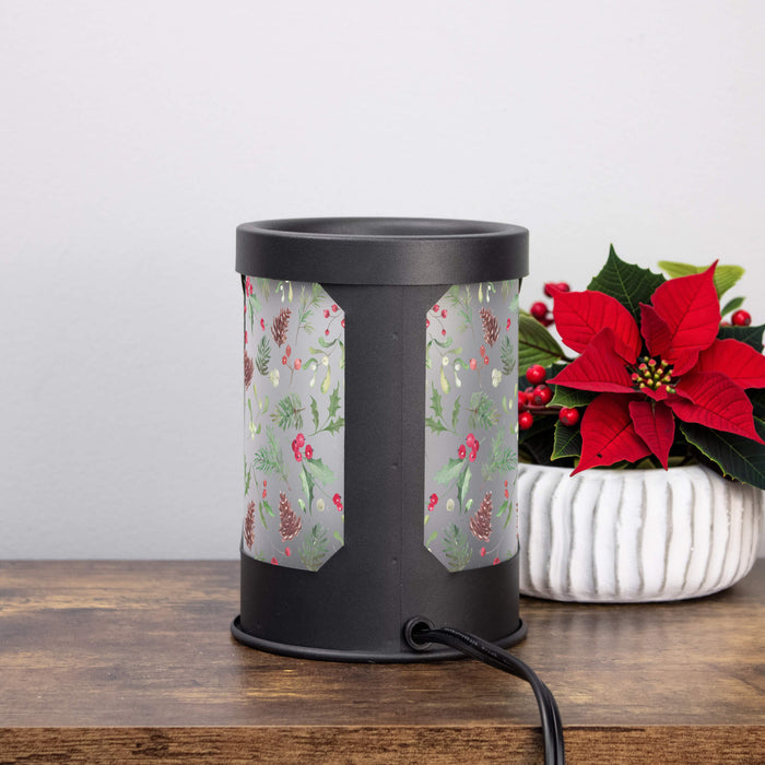 A black cylindrical electric wax warmer with a festive holiday patterned plastic panel, sitting on a wooden table next to a white planter with a red poinsettia.