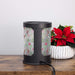 A black cylindrical electric wax warmer with a festive holiday patterned plastic panel, sitting on a wooden table next to a white planter with a red poinsettia.