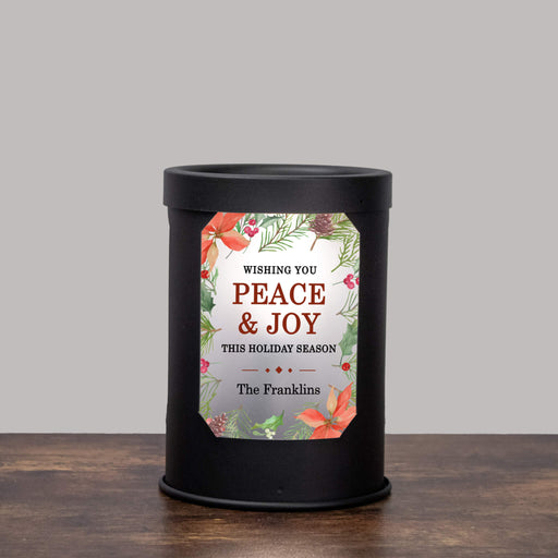 A black cylindrical wax warmer with a decorative holiday label reading 'Wishing You Peace & Joy This Holiday Season The Franklins,' set on a wooden surface against a light gray background.
