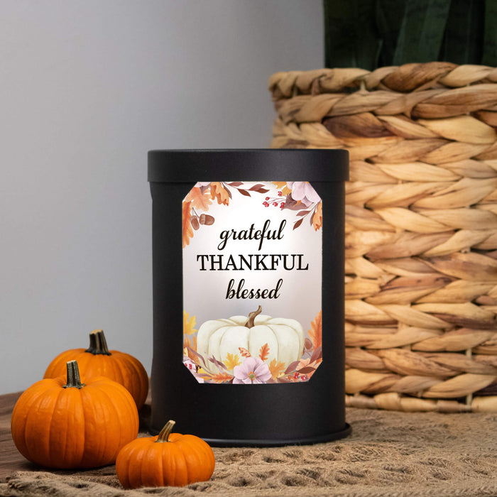 A matte black cylindrical wax warmer with a decorative autumn-themed label reading 'grateful THANKFUL blessed' and a white pumpkin illustration, styled on burlap with small pumpkins and a woven basket in the background