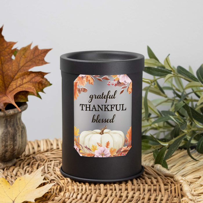 A matte black cylindrical wax warmer with a fall-themed label reading 'grateful THANKFUL blessed' and showing white pumpkins among autumn leaves, styled on a woven mat with leaves and greenery in the background.